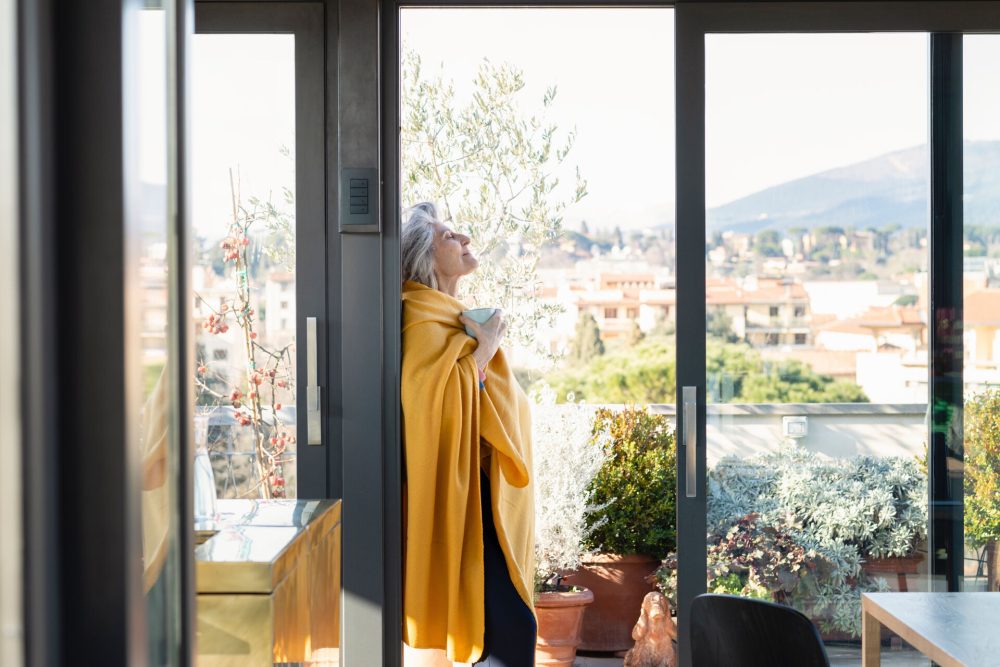 Wohnbedürfnisse der Generation 60+ - In gelbe Decke gehüllte Seniorin steht in geöffneter Balkontür und genießt eine Tasse Kaffe